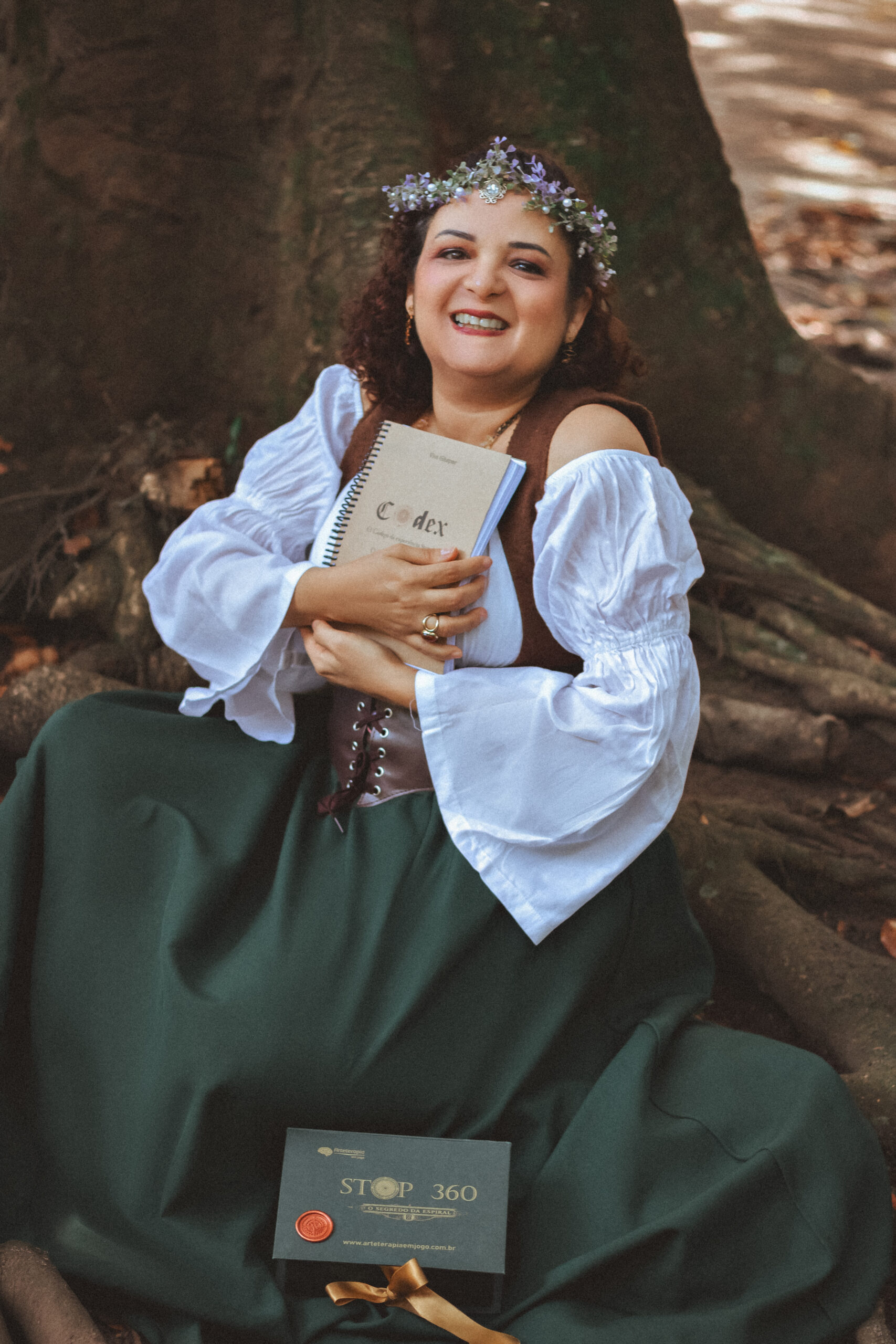 Vivi Gäspar sentada junto a uma árvore, segurando um caderno e o Jogo STOP 360